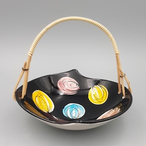 May include: A black ceramic bowl with a glossy finish featuring hand-painted circular motifs in pink, yellow, and blue. The bowl has an irregular, leaf-like rim and is fitted with a curved, light-colored woven rattan handle attached by thin metal wire.
