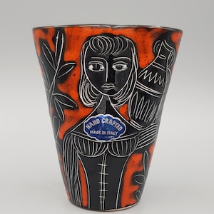 May include: A tapered ceramic vase featuring a vibrant orange and black glaze. The surface displays a sgraffito-style illustration of a figure carrying a vessel, accented by foliage. A blue sticker reads HAND CRAFTED MADE IN ITALY.