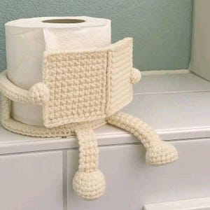 Puede incluir: Un portarrollos de papel higiénico de ganchillo color crema con forma de persona leyendo. El soporte tiene un cuerpo cuadrado, dos brazos y dos piernas con pies redondos. El rollo de papel higiénico es blanco y está dentro del soporte.