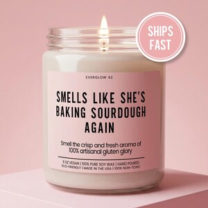 May include: A lit candle in a glass jar with a pink label. The label reads "SMELLS LIKE SHE'S BAKING SOURDOUGH AGAIN." The candle is 9 oz and made with vegan soy wax. A pink circle with "SHIPS FAST" is in the upper right.