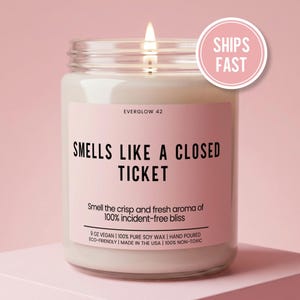 May include: A lit candle in a glass jar with a pink label that reads "SMELLS LIKE A CLOSED TICKET." The label also states "Smell the crisp and fresh aroma of 100% incident-free bliss." The candle is 9 oz and made with soy wax.