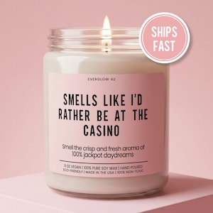 May include: A lit, glass jar candle with a pink label that reads "Smells Like I'd Rather Be At The Casino." The candle is 9 oz and made with soy wax. A pink circle with "Ships Fast" is on the top right.