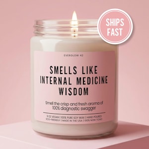 May include: A clear glass jar candle with a lit wick and a pink label. The label reads EVERGLOW 42, SMELLS LIKE INTERNAL MEDICINE WISDOM, Smell the crisp and fresh aroma of 100% diagnostic swagger, 9 OZ VEGAN, 100% PURE SOY WAX, HAND POURED, ECO-FRIENDLY, MADE IN THE USA, 100% NON-TOXIC.