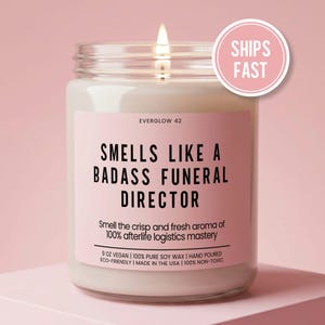 May include: A clear glass jar candle with a white wax fill and a lit wick. The label is light pink with black text reading EVERGLOW 42, SMELLS LIKE A BADASS FUNERAL DIRECTOR, and additional text including 9 OZ VEGAN 100% PURE SOY WAX.