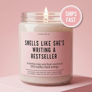 May include: A lit soy wax candle in a clear glass jar with a pink label. The label reads "SMELLS LIKE SHE'S WRITING A BESTSELLER." A pink circle with "SHIPS FAST" is in the upper right corner. The candle is 9 oz.