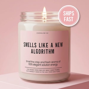May include: A lit candle in a glass jar with a pink label that reads "SMELLS LIKE A NEW ALGORITHM." The label also states "Smell the crisp and fresh aroma of 100% elegant solution energy." The candle is 9 oz and made with soy wax.