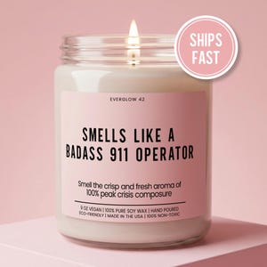 May include: A lit candle in a glass jar with a pink label. The label reads "SMELLS LIKE A BADASS 911 OPERATOR." The candle is made with 9 oz of vegan, 100% pure soy wax, hand-poured, eco-friendly, and non-toxic.