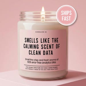 May include: A lit candle in a glass jar with a pink label. The label reads "SMELLS LIKE THE CALMING SCENT OF CLEAN DATA." The jar is 9 oz and made with vegan soy wax. A pink circle with "SHIPS FAST" is in the upper right.