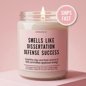 May include: A lit candle in a glass jar with a pink label. The label reads "SMELLS LIKE DISSERTATION DEFENSE SUCCESS." The candle is 9 oz and made with vegan soy wax. A pink circle with "SHIPS FAST" is in the upper right.