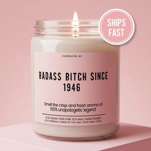 May include: A lit candle in a glass jar with a pink label. The label reads "BADASS BITCH SINCE 1946" and "Smell the crisp and fresh aroma of 100% unapologetic legend." The jar is 9 oz and made with soy wax.