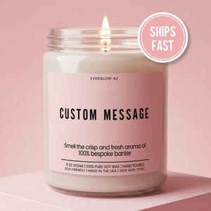 May include: A clear glass jar candle with a lit wick and a light pink label. The label features the text EVERGLOW 42, CUSTOM MESSAGE, and Smell the crisp and fresh aroma of 100% bespoke banter, along with 9 OZ VEGAN, 100% PURE SOY WAX, and HAND POURED.
