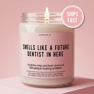May include: A lit candle in a glass jar with a pink label. The label reads "SMELLS LIKE A FUTURE DENTIST IN HERE." The candle is 9 oz, made with vegan soy wax, and hand-poured. A pink circle with "SHIPS FAST" is on the top right.