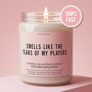 May include: A lit candle in a glass jar with a pink label. The label reads "SMELLS LIKE THE TEARS OF MY PLAYERS." The candle is 9 oz and made with vegan soy wax. A pink circle with "SHIPS FAST" is in the upper right.