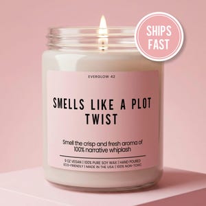 May include: A clear glass candle jar filled with white wax and a lit wick. The label is light pink with black text that reads EVERGLOW 42, SMELLS LIKE A PLOT TWIST, Smell the crisp and fresh aroma of 100% narrative whiplash, 9 OZ VEGAN, 100% PURE SOY WAX.