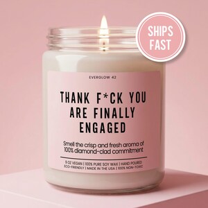 May include: A clear glass jar candle with a lit wick and a pink label. The label reads EVERGLOW 42, THANK F*CK YOU ARE FINALLY ENGAGED, Smell the crisp and fresh aroma of 100% diamond-clad commitment, 9 OZ VEGAN, 100% PURE SOY WAX, HAND POURED.