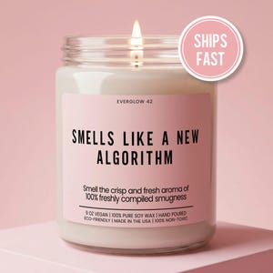 May include: A lit, clear glass jar candle with a pink label that reads "SMELLS LIKE A NEW ALGORITHM." The candle is 9 oz, vegan, and made with soy wax. A pink circle with "SHIPS FAST" is in the upper right corner.