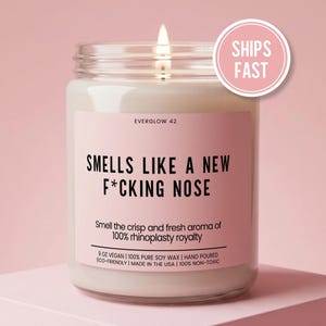 May include: A lit candle in a glass jar with a pink label. The label reads "SMELLS LIKE A NEW F*CKING NOSE" and "Smell the crisp and fresh aroma of 100% rhinoplasty royalty." A pink circle with "SHIPS FAST" is in the upper right.