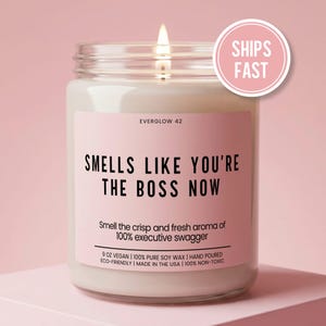 May include: A clear glass jar candle with a lit wick and a light pink label. The label reads: EVERGLOW 42, SMELLS LIKE YOU'RE THE BOSS NOW, Smell the crisp and fresh aroma of 100% executive swagger, 9 OZ VEGAN, 100% PURE SOY WAX, HAND POURED, ECO-FRIENDLY, MADE IN THE USA, 100% NON-TOXIC.