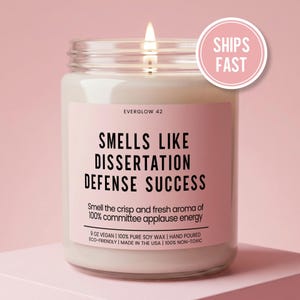 May include: A lit candle in a glass jar with a pink label. The label reads "SMELLS LIKE DISSERTATION DEFENSE SUCCESS." The candle is 9 oz and made with vegan soy wax. A pink circle with "SHIPS FAST" is in the upper right.