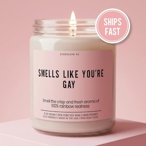 May include: A lit, white candle in a clear glass jar with a pink label. The label reads "SMELLS LIKE YOU'RE GAY" and "Smell the crisp and fresh aroma of 100% rainbow realness." A pink circle with "SHIPS FAST" is in the upper right.