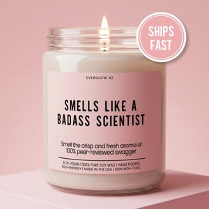 May include: A lit candle in a glass jar with a pink label. The label reads "SMELLS LIKE A BADASS SCIENTIST" and "Smell the crisp and fresh aroma of 100% peer-reviewed swagger." The jar is 9 oz and made with vegan soy wax.