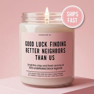 May include: A lit, white candle in a glass jar with a pink label. The label reads "GOOD LUCK FINDING BETTER NEIGHBORS THAN US." The candle is 9 oz, vegan, and made in the USA. A pink circle with "SHIPS FAST" is on the right.