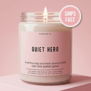 May include: A clear glass jar candle with a lit wick and a pink label. The label reads EVERGLOW 42, QUIET HERO, and includes text about the aroma, 9 OZ VEGAN, 100% PURE SOY WAX, HAND POURED, ECO-FRIENDLY, MADE IN THE USA, and 100% NON-TOXIC.