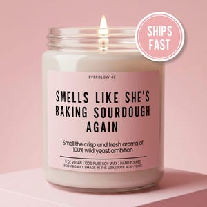 May include: A clear glass jar candle with a lit wick and a light pink label. The label reads: EVERGLOW 42, SMELLS LIKE SHE'S BAKING SOURDOUGH AGAIN, Smell the crisp and fresh aroma of 100% wild yeast ambition, 9 OZ VEGAN, 100% PURE SOY WAX, HAND POURED.