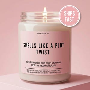 May include: A lit soy wax candle in a glass jar with a pink label that reads "SMELLS LIKE A PLOT TWIST." The candle also says "Smell the crisp and fresh aroma of 100% narrative whiplash." A pink circle with "SHIPS FAST" is on the top right.
