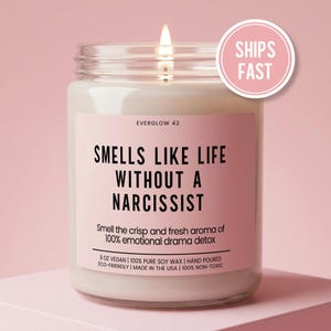 May include: A lit candle in a glass jar with a pink label. The label reads "SMELLS LIKE LIFE WITHOUT A NARCISSIST." The candle is 9 oz and made with soy wax. A pink circle with "SHIPS FAST" is in the upper right.