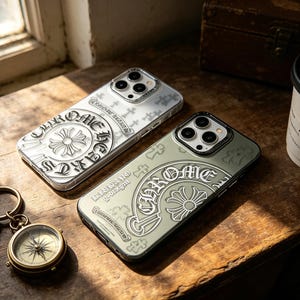 Peut inclure: Deux coques de téléphone avec un motif décoratif et le mot "CHROME" sont présentées sur une surface en bois. Une coque est argentée, l'autre est verte. Une boussole, un porte-clés et une tasse à café sont également visibles.