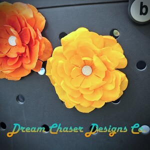 May include: Two fabric flowers, one orange and one yellow, are attached to a black tote bag. The flowers have a sparkly center and are attached to the bag with a button.