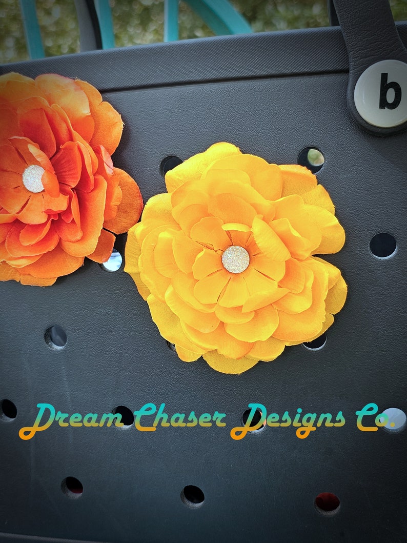 May include: Two fabric flowers, one orange and one yellow, are attached to a black tote bag. The flowers have a sparkly center and are attached to the bag with a button.