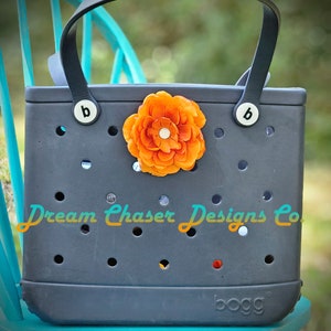 May include: A dark gray tote bag with black leather handles and a bright orange flower embellishment. The bag has a  "Dream Chaser Designs Co." logo and a "bogg" brand name.