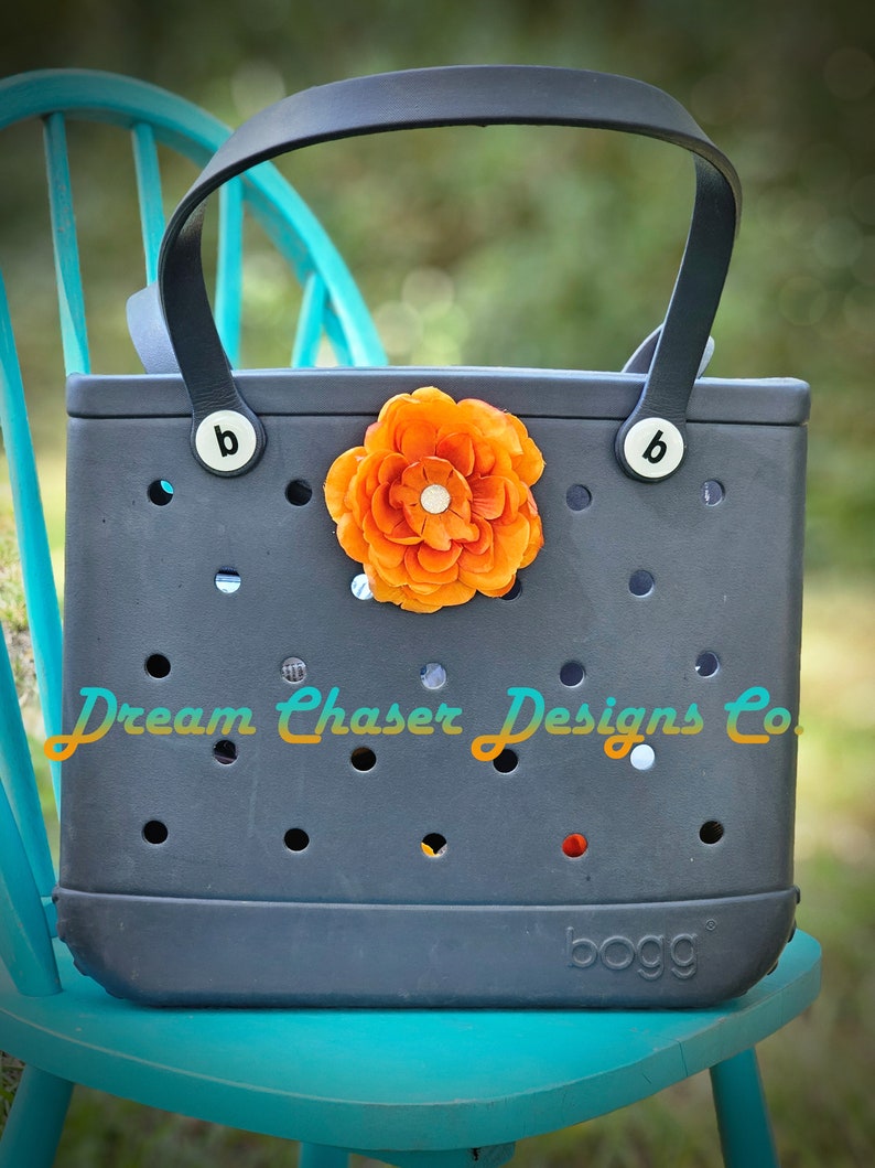 May include: A dark gray tote bag with black leather handles and a bright orange flower embellishment. The bag has a  "Dream Chaser Designs Co." logo and a "bogg" brand name.