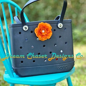 May include: A navy blue Bogg bag with a large orange flower on the front. The bag is sitting on a teal chair. The text "Dream Chaser Designs Co." is printed on the bag.