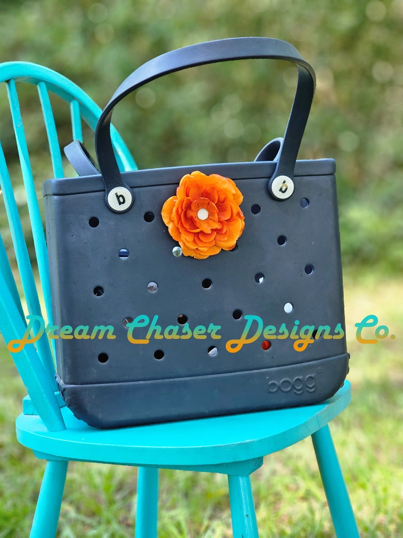 May include: A navy blue Bogg bag with a large orange flower on the front. The bag is sitting on a teal chair. The text "Dream Chaser Designs Co." is printed on the bag.