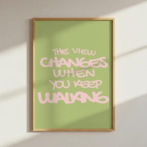 Op de afbeelding: Een rechthoekige wandkunstprint in een lichthouten lijst met een saliegroene achtergrond en de tekst THE VIEW CHANGES WHEN YOU KEEP WALKING in een vetgedrukt, handgetekend lichtroze lettertype in het midden van de print.
