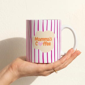 Op de afbeelding: Een witte keramische mok met verticale magentakleurige strepen en een beige, organisch gevormde vlek in het midden. De tekst Mamma's Coffee is in oranje letters gedrukt, waarbij een klein roze hartje de letter o in Coffee vervangt.