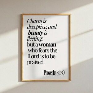 May include: A framed wall art print featuring black serif typography on a white background. The text reads: Charm is deceptive, and beauty is fleeting; but a woman who fears the Lord is to be praised. Proverbs 31:30. The frame is light wood.