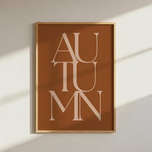 May include: A rectangular wall art print in a light wood frame featuring a deep terracotta brown background with the word AUTUMN printed in large, light beige serif capital letters stacked vertically in the center of the composition.