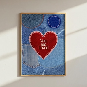 Puede incluir: Una impresión de arte mural rectangular que presenta un collage de parches de tela de mezclilla azul con costuras visibles. Un gran corazón rojo central con un borde festoneado y costuras blancas contiene el texto blanco You are Loved.