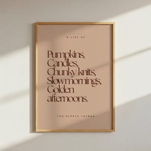 May include: A rectangular wall art print in a light wood frame featuring a tan background with dark brown text that reads: A LIST OF Pumpkins, Candles, Chunky knits, Slow mornings, Golden afternoons. THE SIMPLE THINGS, centred in a clean layout.