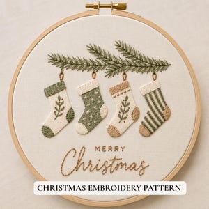 May include: A round wooden embroidery hoop containing white fabric with a hand-stitched design of four hanging Christmas stockings on pine branches. The stockings feature green and tan patterns. The text Merry Christmas is stitched below.