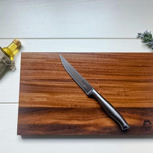 Op de afbeelding: Een rechthoekige houten snijplank met een gekarteld mes van roestvrij staal. Een kleine fles olie in een jute zak en een takje lavendel zijn ook aanwezig. De snijplank heeft een donkerbruine afwerking.