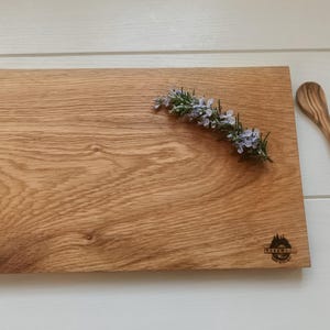 Op de afbeelding: Een rechthoekige houten snijplank met een takje rozemarijn en een kleine houten lepel. De plank heeft een natuurlijke houtnerf en een logo. De lepel is ongeveer 15 cm lang.
