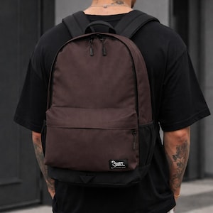 Puede incluir: Una mochila marrón con correas negras y un bolsillo frontal. La mochila tiene un diseño sencillo con la palabra "Start" impresa en una pequeña etiqueta. La mochila la lleva una persona.
