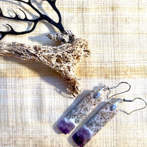May include: A pair of handmade earrings with a rectangular shape, featuring a speckled, light-colored top and a vibrant purple bottom. The earrings hang from silver-colored fish hook ear wires. The background includes a piece of driftwood and a dark, branch-like object.