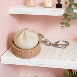 Puede incluir: Un llavero beige con forma de dumpling con un soporte marrón en forma de cesta. El dumpling tiene pliegues detallados y el soporte tiene una textura tejida. Se adjunta una cadena y un llavero plateados.