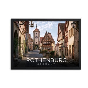Op de afbeelding: Een ingelijste kunstdruk met een straatbeeld in Rothenburg, Duitsland, met vakwerkhuizen en een klokkentoren. De tekst ROTHENBURG GERMANY staat in witte hoofdletters onderaan in het midden van de afbeelding gedrukt.